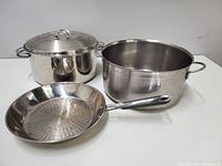 Photo showing the front view of the stainless steel frying pan and two large pots, one with lid and one without, all placed on a flat surface.