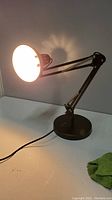 Image of black adjustable table lamp turned on, showing light illumination from lamp head and articulating arm.