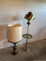 Two old-style brass floor lamps placed next to a cinder block wall. One has a dome brass shade and mid-pole tray, the other has a white cylindrical shade.