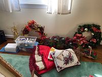 Photo showing assortment of Christmas wreaths decorated with greenery, red berries, pinecones, and large red bows arrayed along the floor beneath a window. Red and cream fabric pieces and two decorative Christmas pillows with poinsettia designs next to the wreaths. Clear plastic bin and white boxes filled with Christmas ornaments on the left side.