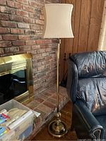 Brass floor lamp with beige curved fabric shade beside a brick fireplace and leather chair.