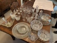 Wide view of assorted clear crystal and glass serving pieces, bowls, trays, and various glasses arranged on a wooden table.