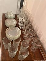 Photo showing 27 various glasses including tumblers and stemmed glasses, stacked clear glass plates, and a clear soup bowl on a wooden surface.