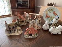Seven detailed miniature Scottish cottages displayed on a wooden table, each with distinct roofs, chimneys, and detailed architecture.