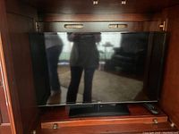 Photo of a black flat screen JVC television with remote control inside a wooden cabinet, screen reflecting background and people.