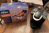Photo showing Keurig coffee maker and boxed Oster immersion deep fryer on wooden table.