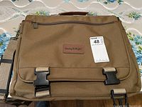 Front view of tan canvas laptop bag showing the flap with two black quick-release buckles, zippered pocket, and brand label patch 'Doing It Right'.