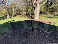 Photo showing a petite black iron outdoor furniture set including one loveseat, three chairs, and one small round table arranged outdoors on a dirt/rock surface with trees and grass in the background.