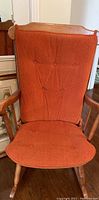 Photo showing the orange cushioned wooden rocking chair from a front angle, highlighting the cushioned seat and backrest, button tufting and light oak wooden armrests and legs.