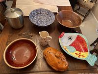 Photo showing multiple items: silver-tone metal ice bucket with lid, blue and white patterned ceramic bowl, natural wood bowl, small speckled pitcher with blue polka dots, fish-shaped ceramic serving plate with colorful design, reddish-brown ceramic covered dish, and small kitchen accessories including scissors and measuring spoon.
