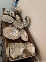 Wide view of multiple large silver plated serving trays and bowls arranged on a wooden sideboard.