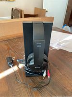 Black Sony wireless headphones placed on black vertical charging stand on a wooden table. Charging cable with plug and RCA connectors coiled around stand base.