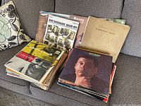 Two stacks of vinyl record albums on a gray couch showcasing a variety of genres and artists.