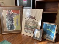 Four framed artworks leaning against a wooden shelf and a TV stand on hardwood floor.