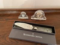 Photo displays two Waterford crystal desk clocks and one Waterford crystal cake server in original box on wooden surface.