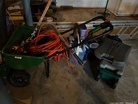 Wide shot of wheelbarrow loaded with orange extension cords and hand tools, green electric tool, and miscellaneous yard equipment.