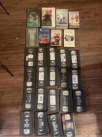 27 VHS tapes laid out on floor, some with handwritten or printed labels, no covers included. Some recognizable titles on printed labels.