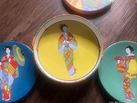 Top view of three plates showing colorful Japanese Geisha illustrations on solid yellow, blue, and green backgrounds.