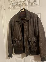Dark brown leather bomber jacket with button front and ribbed cuffs and hem, hanging on a white door rack.