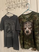 Two large t-shirts hanging on hangers: one grey with 'Snake Bite Rhythm & Blues' print and one camouflage with a bear wearing sunglasses graphic.