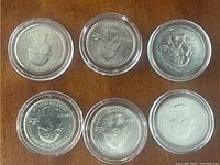 Six U.S. quarter coins each in a clear protective round plastic container displayed on wooden surface.