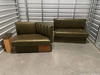 Two greenish RV sofa sections placed side by side in a storage unit, showing the upholstery, foam cushions, low backs, wooden base with damage on one piece.
