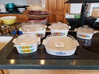 Photo showing six pieces of vintage glass bakeware with floral decoration and clear glass lids placed on a black stovetop surface.