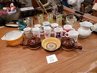 Photo showing mix of various coffee mugs, glasses, decorative bowls and a white ceramic pitcher on a wooden table.