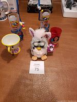 Photo showing four Disney plastic cups including a yellow chip mug and a white Furby toy with glasses and pink ears on a wooden surface.