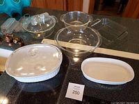 Full lot view on kitchen counter showing two CorningWare covered white ceramic dishes, clear glass Pyrex casserole with lid, two Anchor Hocking clear glass bowls, and two white ceramic baking dishes.
