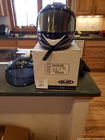 Front view of HJC helmet on box showing the helmet's red, black, white graphic design and tinted visor. Additional clear visor next to box.