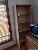 Single image of a wooden bookshelf with three shelves placed next to a window and a dresser with a TV on top.