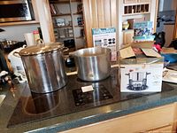 Large and small stainless steel soup pots on cooktop along with boxed stainless steel fondue set