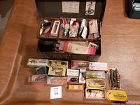Open metal tackle box containing vintage fishing lures, boxes, and spools inside on a leather surface.