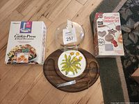 Photo showing wooden cheese/serving platter with glass dome cover, decorative floral plate on platter, two boxed cookie press and rosette sets, and partial view of wicker basket on carpet.