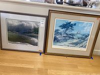 Photo of two framed artworks side by side on the floor, one with waves and foam and the other a sailing ship scene.