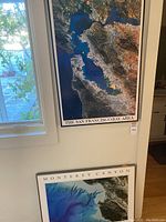 Photo showing both framed maps side by side near window: San Francisco Bay Area satellite photo on wall and Monterey Canyon seafloor map below it on the floor leaning against wall.