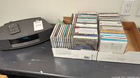 Bose Wave Radio/CD player with digital clock display, remote on top, and box of assorted CDs next to it on a surface.