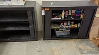 Front view of two metal cabinets with black plastic rolling doors; right cabinet open showing multiple shelves with paint bottles and model painting supplies.