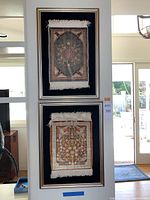Pair of small framed carpets mounted on black velvet, shown hanging vertically on a white wall.
