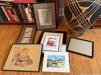 Five framed art pieces and one empty frame arranged on wood floor near bookcase and large ceramic vase.