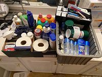 Three boxes with assorted bathroom supplies including body powder, shaving cream, shampoo bottles, and toilet paper, shown on a bathroom counter.