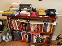 Four shelves with numerous books of varying sizes and colors, stacked both vertically and horizontally, along with two rotary dial telephones and a small decorative item on top.