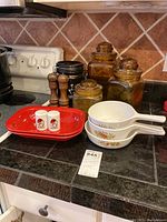 Overall kitchen lot photo showing CorningWare pans, amber canisters, salt and pepper shakers, grinders, and black bowls on countertop.