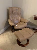 Photo of tan leather recliner chair and matching footstool in corner, showing overall shape and condition, scratches and stains visible on chair headrest.