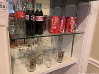 Shelf displaying Coca-Cola glass bottles including original flair recreations, classic full glass bottles, and branded drinking glasses beneath cans.
