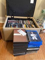 Stacked DVDs and Blu-ray discs with visible titles and beige storage ottoman containing additional DVDs and CDs.