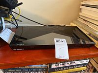 Sony Blu-ray/DVD player with a glossy black surface on a wooden shelf, surrounded by books