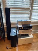 Sony sound bar and speakers with HP printer and cartridge boxes stacked beside them on wood floor near window.
