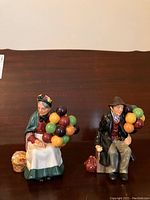 Photo of two seated porcelain or ceramic figurines, a man and a woman, holding clusters of colorful spherical objects, with a basket and sack beside them.